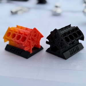 Könnte beinhalten: Zwei 3D-gedruckte Miniatur-Motorblöcke auf schwarzen rechteckigen Sockeln. Ein Motorblock zeigt einen leuchtenden orange-gelben Farbverlauf, während der andere einfarbig schwarz ist. Beide Modelle haben das Wort HEMI auf dem vorderen Sockel eingeprägt.