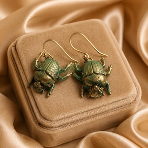 May include: A pair of dangle earrings featuring detailed beetle charms with a weathered green and gold finish. The earrings are designed with simple gold-toned metal hooks for attachment, showcasing a textured, insect-inspired aesthetic.