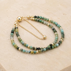 May include: A double-strand bracelet featuring small, faceted green and teal gemstone beads interspersed with polished gold-tone metal spheres. The bracelet includes a gold-tone sliding clasp mechanism with a delicate chain for adjustment.