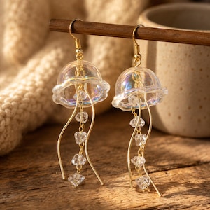 Può includere: Un paio di orecchini pendenti con ciondoli a forma di medusa iridescente a cupola. Ogni orecchino include ganci in metallo dorato, sottili fili d'oro curvi e una catenella centrale decorata con piccole perline di cristallo irregolari.
