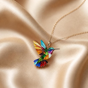 May include: A gold-toned necklace featuring a pendant shaped like a hummingbird. The bird is decorated with vibrant, multi-coloured enamel sections in shades of blue, green, yellow, orange, and red, separated by thin gold-coloured metal lines.