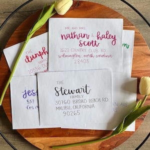 May include: A collection of white envelopes featuring various custom calligraphy addresses in black, purple, and magenta ink, including names like Nathan & Haley Scott and The Stewart Family, accented by two pale yellow tulip stems.