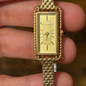 May include: A gold-tone rectangular Accutron wristwatch featuring a textured link band and a rope-style bezel. The gold dial displays the text Accutron, Swiss, and Quartz 9409, with thin gold hour markers and a small black crown on the right side.