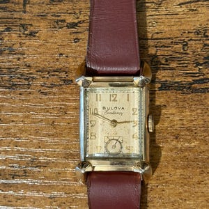 May include: A vintage Bulova Excellency rectangular wristwatch with a gold-tone case, a cream-colored dial featuring gold Arabic numerals, and a dark maroon leather strap. The watch face displays the Bulova brand name and Excellency script.