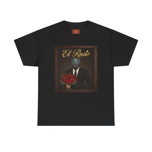 Puede incluir: Una camiseta negra de manga corta con un estampado gráfico de una persona con máscara de lucha azul y traje negro, sosteniendo un ramo de rosas rojas. El texto "El Rudo" está impreso en dorado sobre la figura, con un logo "RUDO" en el cuello.