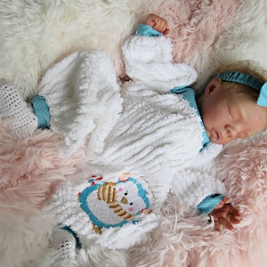 以下が含まれることがあります： 青いペンギンのデザインと青いリボン付きヘッドバンドが付いた白いベビー服。この服は柔らかくふわふわした素材で作られており、新生児に最適です。