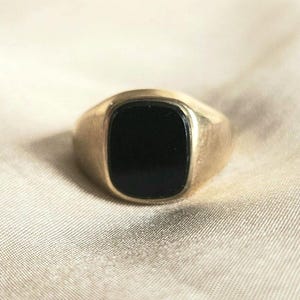 May include: A gold-toned signet ring featuring a smooth, rectangular black onyx inlay with rounded corners. The metal band has a polished finish and a solid, tapered design that frames the central black stone securely within the setting.