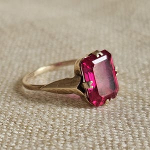 May include: A gold ring featuring a large, rectangular, emerald-cut vibrant pink gemstone held in place by four prongs. The band has a pointed, triangular shoulder design that connects to the setting, showcasing a polished metallic finish.