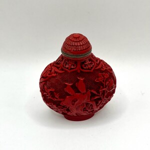 May include: A small, round, bright red snuff bottle featuring intricate, deeply carved relief patterns of birds, foliage, and geometric lattice designs. The bottle is topped with a matching red, textured, dome-shaped lid with a thin metal rim.