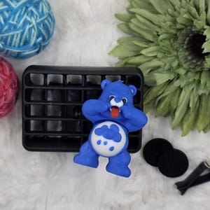 May include: A blue Care Bear figurine with its tongue sticking out, positioned next to a black vent clip. The bear has a cloud design on its belly. Also included are two black felt pads, a black vent clip, and two balls of yarn.