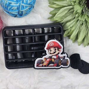 May include: A black plastic car vent with a Mario Kart character sticker. The sticker features Mario in a red racing suit driving a red race car.