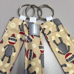 May include: Three keychains with a beige fabric background and a pattern of gray monkeys wearing red and white hats. The keychains have silver key rings and white fabric loops.