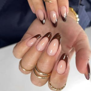 Pode incluir: Unhas em formato de amêndoa com uma base nude brilhante e pontas francesas castanho-escuras. Os dedos estão adornados com anéis finos de metal dourado polido no dedo médio e anelar, complementando a estética cuidada da manicure.