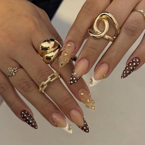 May include: A collection of gold-toned rings including a thick knotted band, a chain-link band, a floral-shaped ring, and a smooth domed ring. The fingernails are shaped into long, sharp points with brown and white polka dot patterns and gold accents.
