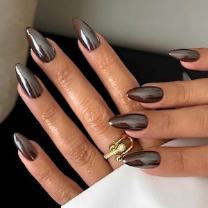 May include: Almond-shaped fingernails with a glossy, dark brown chrome finish. A gold-toned ring with a textured, cylindrical bar design and small embedded clear stones sits on the ring finger, featuring a double-banded open structure.