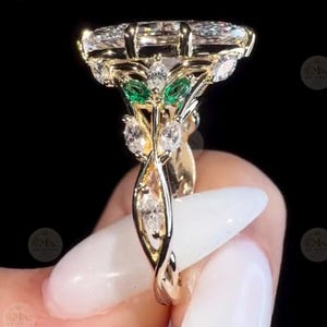 May include: A gold ring featuring a large marquise-cut center stone, accented by smaller marquise-cut clear stones and two vibrant green marquise-cut stones set in a leaf-like pattern along the band, showcasing intricate metalwork and prong settings.