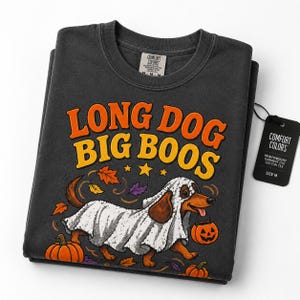Può includere: Una t-shirt girocollo grigio antracite con la grafica di un bassotto vestito da fantasma bianco, che tiene in mano un secchiello a forma di zucca. Il testo recita LONG DOG BIG BOOS in arancione e giallo. Include un'etichetta nera Comfort Colors.