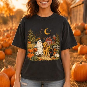 Peut inclure: Un t-shirt gris anthracite avec une impression graphique représentant un fantôme portant un chapeau de sorcière, un golden retriever et un chat noir marchant dans un champ de citrouilles près d'une grange rouge sous un croissant de lune.