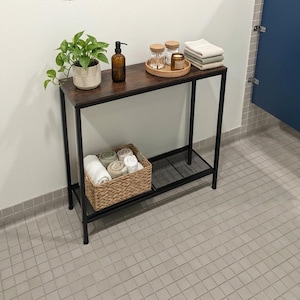 May include: A rectangular console table with a dark wood-grain top and a black metal frame. The top holds a potted green plant, a brown soap dispenser, and a round tray with two jars and folded towels. A woven basket with towels sits on the lower shelf.