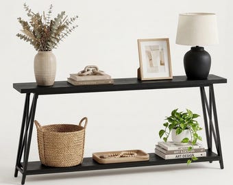 Könnte beinhalten: Ein schwarzer Konsolentisch mit X-förmigem Untergestell und unterer Ablage. Oben stehen eine beige Vase mit getrocknetem Eukalyptus, Bücher mit Holzkette, ein Bilderrahmen und eine schwarze Lampe. Unten befinden sich ein geflochtener Korb, ein Holztablett mit Perlen und eine Topfpflanze auf Büchern.