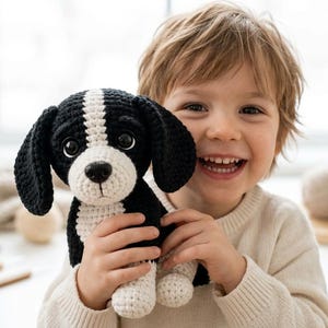 Pode incluir: Um brinquedo de pelúcia feito à mão em crochê, com fios pretos e brancos, grandes olhos de plástico escuro e orelhas caídas. O animal tem o focinho e uma mancha no peito brancos, com a cabeça, orelhas e costas na cor preta, em posição sentada.