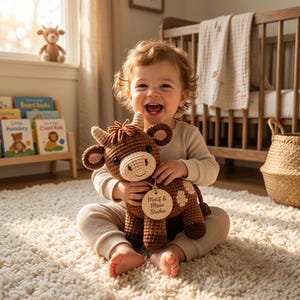May include: A brown and cream crocheted cow stuffed animal with a round wooden tag hanging from its neck that reads Motif & Muse Studio. The toy features small horns, dark eyes, and a textured yarn finish, held in a seated position.