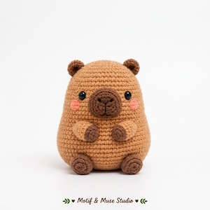 Peut inclure: Une peluche capybara en crochet beige avec un museau marron foncé, de petites oreilles arrondies et des yeux en perles noires. La figurine présente des joues roses brodées, des pattes et des pieds marron foncé. Texte Motif & Muse Studio.