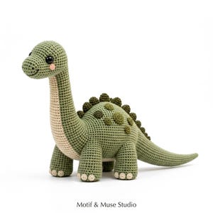 Peut inclure: Un jouet dinosaure au crochet dans des tons vert sauge et crème. La figurine présente un long cou, un corps arrondi avec des taches circulaires vert foncé, une rangée de petites pointes sur le dos et des yeux de sécurité noirs. Le texte Motif & Muse Studio est visible.