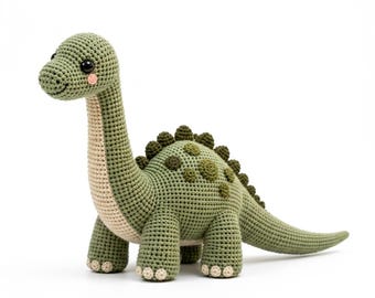 May include: A crocheted dinosaur toy in shades of sage green and cream. The figure features a long neck, a rounded body with dark green circular patches, a row of small spikes along the back, and black safety eyes. The text Motif & Muse Studio is visible.