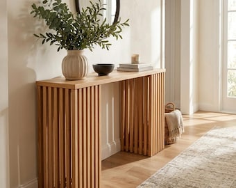 Puede incluir: Una mesa consola de madera clara con dos bases de listones verticales. Encima hay un jarrón de cerámica beige acanalado con ramas de eucalipto verdes, un cuenco negro pequeño y una pila de libros con una vela encima.