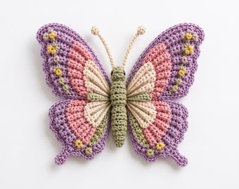 Puede incluir: Una mariposa de ganchillo con un cuerpo central verde oliva y dos antenas. Las alas están superpuestas con hilo morado, rosa y crema, acentuadas por pequeños detalles florales amarillos a lo largo de los bordes exteriores de las alas moradas.