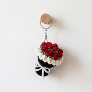 May include: A crochet keyring featuring a miniature bouquet of red roses with green leaves, wrapped in black fabric with a white ruffled trim and a white tied ribbon. The bouquet is attached to a silver metal keyring and chain.