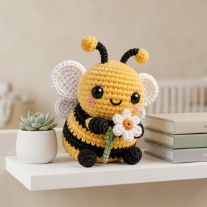 May include: A crocheted bee plush toy with a yellow and black striped body, white wings, and black antennae. The bee has large black eyes, pink cheeks, and a smiling mouth, and it is holding a small white flower with a yellow center in its hands.