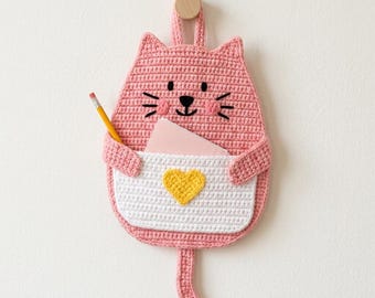 Puede incluir: Un organizador de pared de ganchillo con forma de gato rosa, con un bolsillo blanco, un corazón amarillo, rasgos faciales bordados en negro y una cola. Un lápiz amarillo y una nota rosa pálido están dentro del bolsillo, colgado de un gancho.