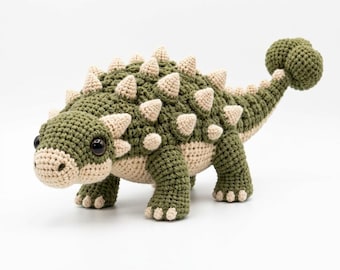 May include: A crocheted ankylosaurus toy featuring a dark olive green body with cream-colored spikes along its back and tail. The dinosaur has large dark brown eyes, a cream-colored snout, and a clubbed tail. The text Motif & Muse Studio is visible.