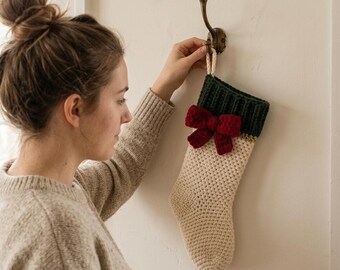 May include: A cream-colored crocheted Christmas stocking featuring a dark green ribbed cuff and a prominent deep red crocheted bow attached to the front. The stocking is suspended from a brass-toned metal wall hook.
