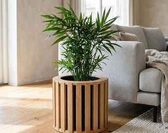 Puede incluir: Una planta de palmera de salón verde en una maceta cilíndrica de madera. La maceta presenta un diseño de listones con tiras verticales de madera de roble de color marrón claro dispuestas alrededor de un recipiente interior oscuro.
