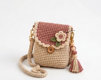Puede incluir: Un bolso bandolera de ganchillo con cuerpo beige y solapa rosa empolvado con borde festoneado en color crema. La solapa está decorada con una pequeña flor de ganchillo y dos hojas verdes. Un botón de madera cierra la solapa y una borla con cuentas de madera cuelga del lateral.