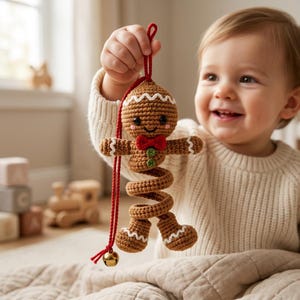 May include: A brown crocheted gingerbread person ornament with a spiral body, a red bow tie, two green buttons, and white icing details. The ornament features a red hanging cord with a small gold-colored metal bell attached at the bottom end.