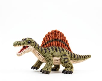 Puede incluir: Un juguete de dinosaurio Spinosaurus tejido a ganchillo con cuerpo verde y rayas marrón oscuro, vientre color crema y una prominente vela dorsal de color naranja amarronado. La figura tiene ojos negros, boca abierta con dientes blancos y garras oscuras.