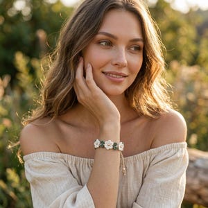 May include: A bracelet featuring a series of alternating white and dark green crocheted flower motifs. The floral elements are connected in a row, with a small gold-toned bead centered in each white flower. The bracelet includes a thin cord tie.