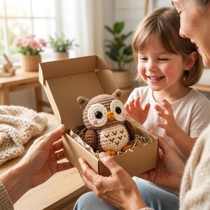 May include: A brown and cream crocheted owl toy with large black eyes, a small orange beak, and pink cheeks, nestled inside a brown cardboard gift box filled with shredded paper filler, presented as a gift for a young child to enjoy.
