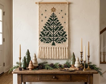 May include: A cream-colored crocheted wall hanging featuring a dark green Christmas tree pattern with a gold star at the top, finished with a fringe at the bottom and suspended from a wooden dowel with a string.