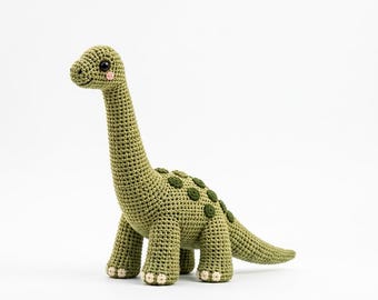 Puede incluir: Un juguete de dinosaurio tejido a ganchillo en color verde oliva claro con cuello y cola largos. El cuerpo presenta manchas circulares de color verde oscuro en la espalda, pequeños ojos de seguridad negros y mejillas rosadas. El texto Motif & Muse Studio es visible.