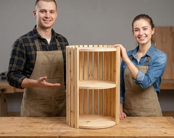 Puede incluir: Una estantería cilíndrica de madera con tres niveles circulares conectados por listones verticales. La estructura está hecha de madera natural clara con veta visible, mostrando un diseño abierto con un marco exterior de listones.