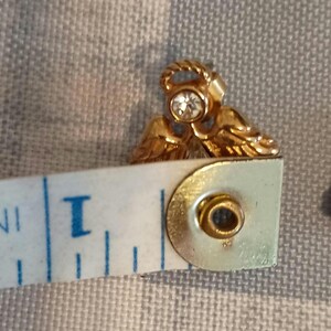 May include: A gold-colored angel-shaped earring with a clear, round gemstone in the center. The earring is approximately 0.75 inches in length. The angel has detailed wings and a halo.
