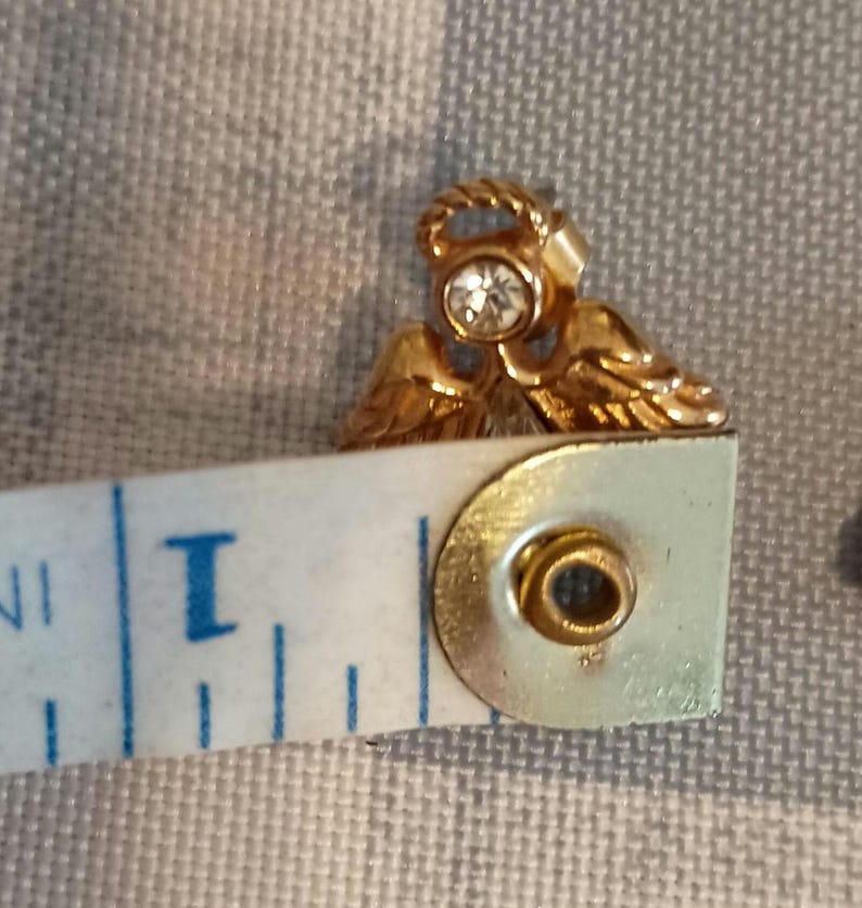 May include: A gold-colored angel-shaped earring with a clear, round gemstone in the center. The earring is approximately 0.75 inches in length. The angel has detailed wings and a halo.