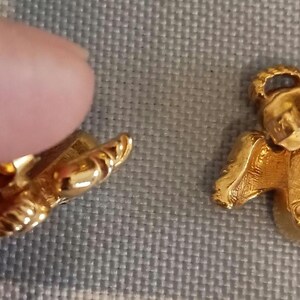 May include: Close-up of two gold-colored angel-shaped earrings. The earrings have detailed wings and a textured surface. One earring is partially obscured by a finger. The earrings are on a gray surface.