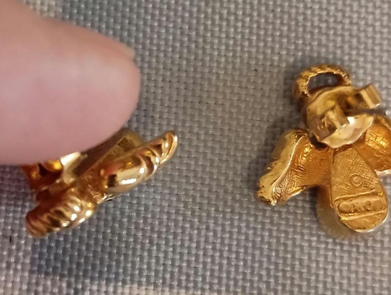 May include: Close-up of two gold-colored angel-shaped earrings. The earrings have detailed wings and a textured surface. One earring is partially obscured by a finger. The earrings are on a gray surface.