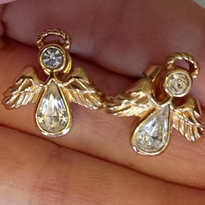 May include: A pair of gold-toned angel earrings. Each earring features a clear, teardrop-shaped crystal pendant and a smaller, round crystal at the head. The angels have detailed wings and halo accents.